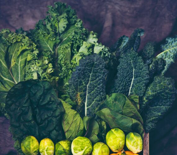 A colorful arrangement of cruciferous vegetables including dark green curly kale, lacinato kale (dinosaur kale), Swiss chard, and fresh Brussels sprouts, all displayed in a rustic wooden crate against a moody purple fabric background.