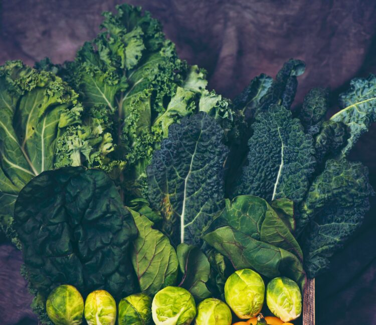 A colorful arrangement of cruciferous vegetables including dark green curly kale, lacinato kale (dinosaur kale), Swiss chard, and fresh Brussels sprouts, all displayed in a rustic wooden crate against a moody purple fabric background.