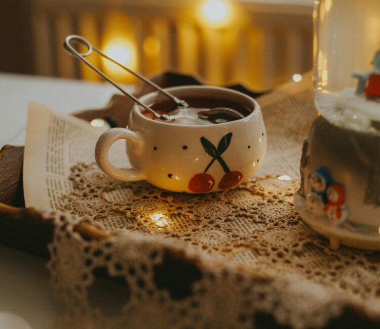 Hands lifting the lid of a clear glass teapot filled with amber herbal tea, surrounded by small bowls of dried herbs, honey, and empty glass teacups on a wooden board in soft natural light.