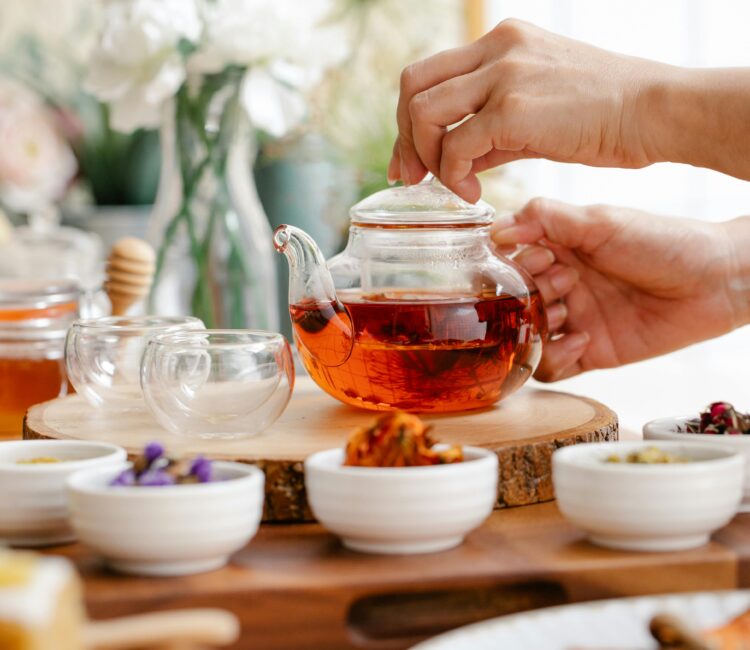 Hands preparing a pot of herbal tea surrounded by dried herbs and cups on a wooden table