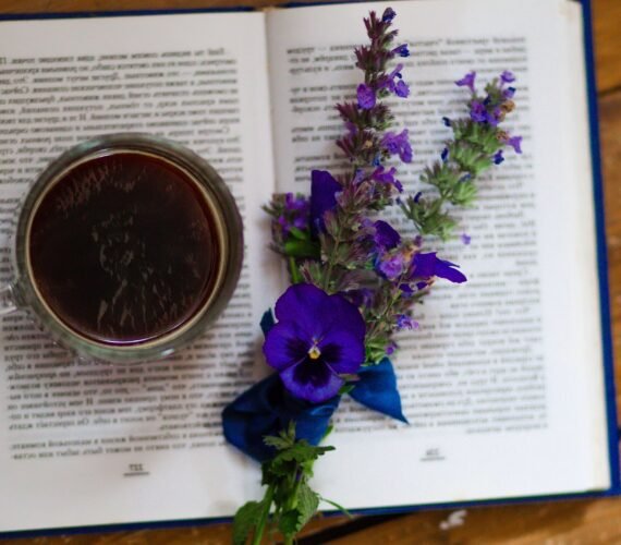 A glass mug of dark herbal tea resting on an open book, decorated with purple and blue wildflowers arranged across the pages on a rustic wooden table.