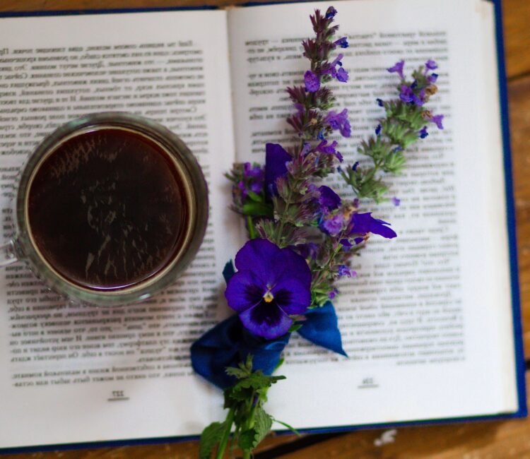A glass mug of dark herbal tea resting on an open book, decorated with purple and blue wildflowers arranged across the pages on a rustic wooden table.
